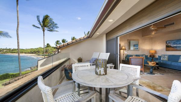 A beachfront patio with white furniture overlooks the ocean, palm trees sway, and an adjacent cozy indoor living space adorned with blue accents.