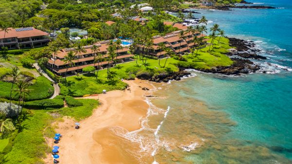 This image shows a tropical beach with clear waters, lush greenery, resort buildings, and a few people and umbrellas on the sand.