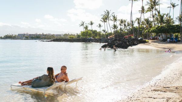 Two people relax on a floating lounge chair in a serene beach setting, surrounded by palm trees and gentle waves under a partly cloudy sky.