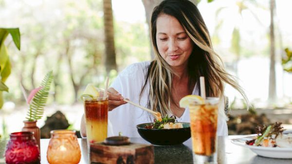 A woman is enjoying a meal at an outdoor table, surrounded by drinks and food, with trees visible in the background.