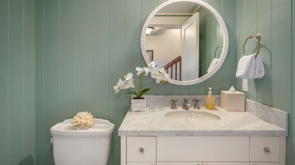 The image shows a bathroom with light green vertical panel walls, a round mirror, a sink with a white countertop, and a toilet with a decorative shell.