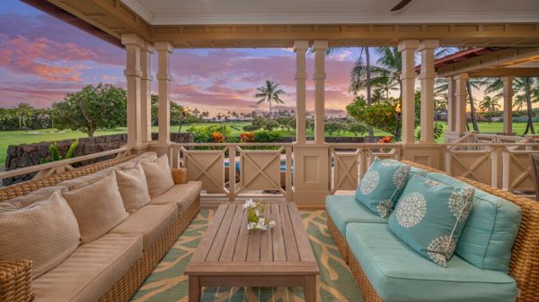 This image features a cozy outdoor lounge area with wicker furniture, colorful cushions, and a wooden table, overlooking a tropical landscape and sunset.