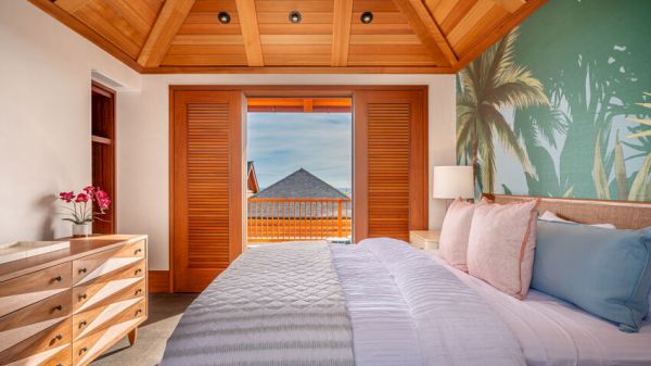 The image shows a tropical-themed bedroom with a wooden ceiling, a double bed, a dresser with flowers, and an open door leading to a balcony with a sea view.