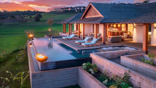 A luxurious villa with an infinity pool, lounge chairs, and fire features overlooking a scenic landscape, set against a beautiful sunset sky.