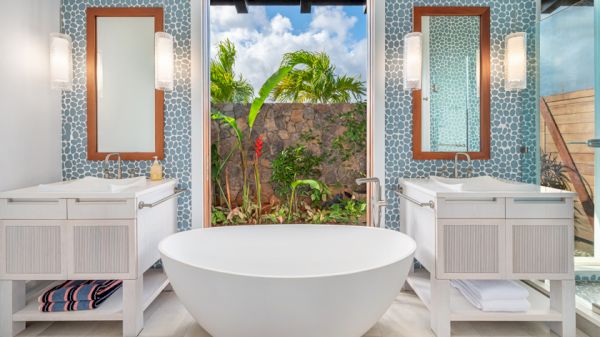 Modern bathroom with a standalone tub, twin sinks, mirrors, and windows revealing a lush, tropical garden view.