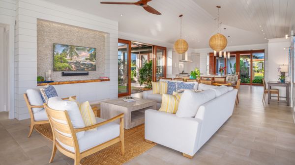 A spacious, modern living room with white sofas, wooden furniture, a mounted TV, ceiling fans, and large windows leading to an outdoor area.