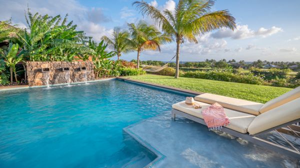 The image shows a serene pool with waterfalls, surrounded by lush greenery and palm trees, along with a comfortable lounge chair under a clear sky.