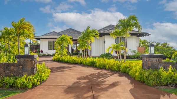 A driveway leading to a white single-story house surrounded by lush greenery and palm trees, with the address number "3555" visible on a stone pillar.