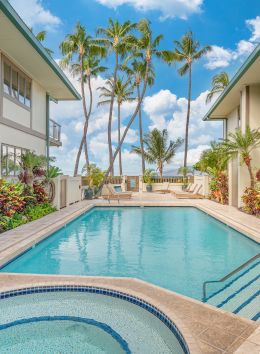 The image shows a serene courtyard with a pool and hot tub, surrounded by tropical plants and two-story buildings, with palm trees and blue skies.