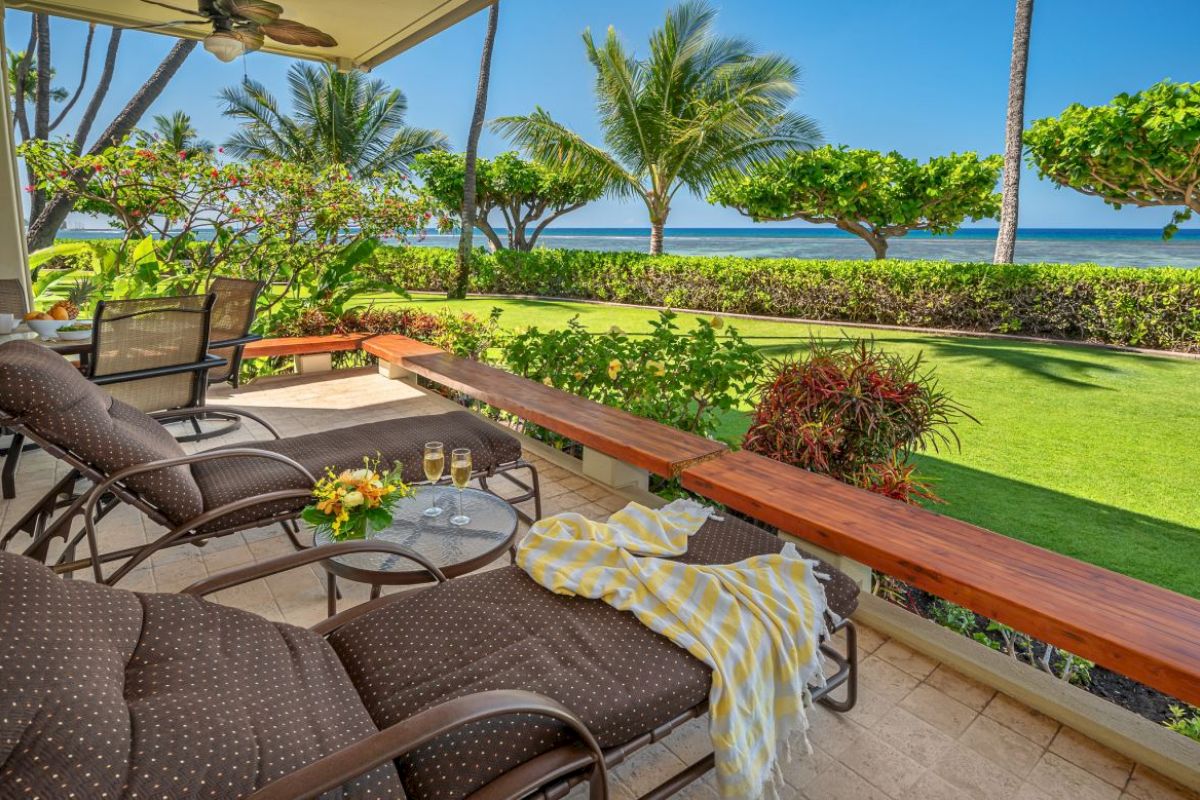 A relaxing patio with lounge chairs, a table with glasses and a flower arrangement, overlooking a garden with palm trees and the ocean in the background.