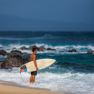 A person holding a surfboard and standing on a beach, looking at the ocean waves.