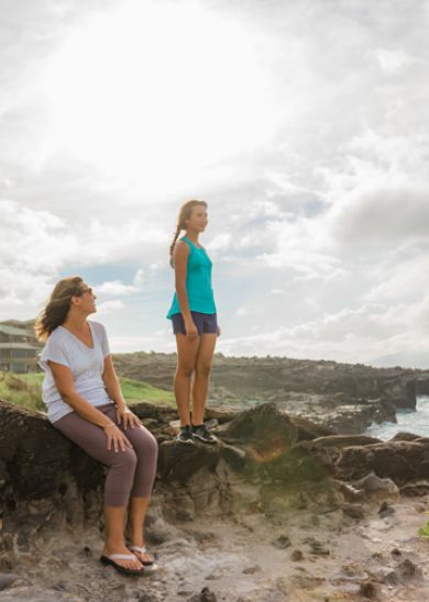 Two people are on a rocky coastline; one is seated while the other stands, gazing out at the ocean under a cloudy sky.