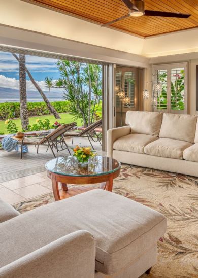 A bright living room with beige sofas, a wooden coffee table, and a large rug, opening to a patio with chairs and a scenic view of the ocean.