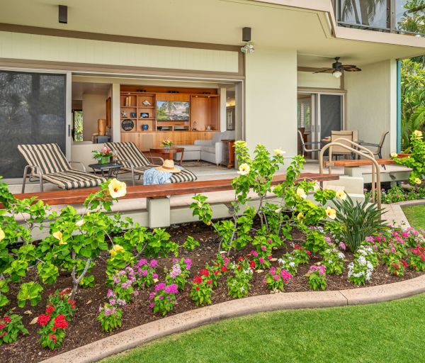 Outdoor patio with lounge chairs, a table, and flowers in front of a modern house with sliding doors opening to an indoor living space.