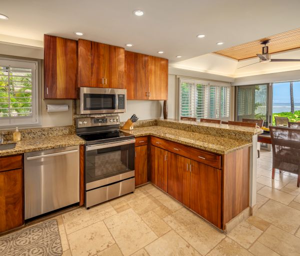 A modern kitchen with stainless steel appliances, wooden cabinets, an island, and a view of the dining area and ocean through glass doors.