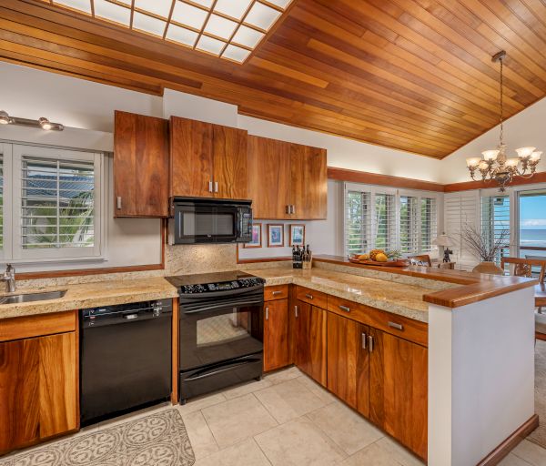 A modern wooden kitchen with black appliances, light countertops, and a dining area with a chandelier and large windows offering an ocean view.