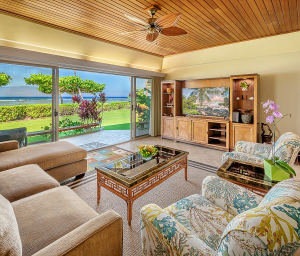 A cozy living room with large sliding doors opening to a lush garden view. It features a sectional sofa, floral chairs, and a TV on a wooden cabinet.