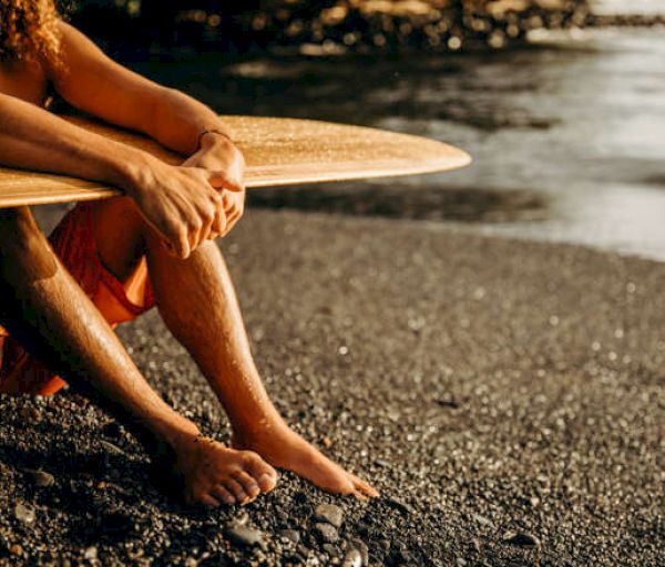 A person sits on the shore with a surfboard resting on their knees, waiting by the water.