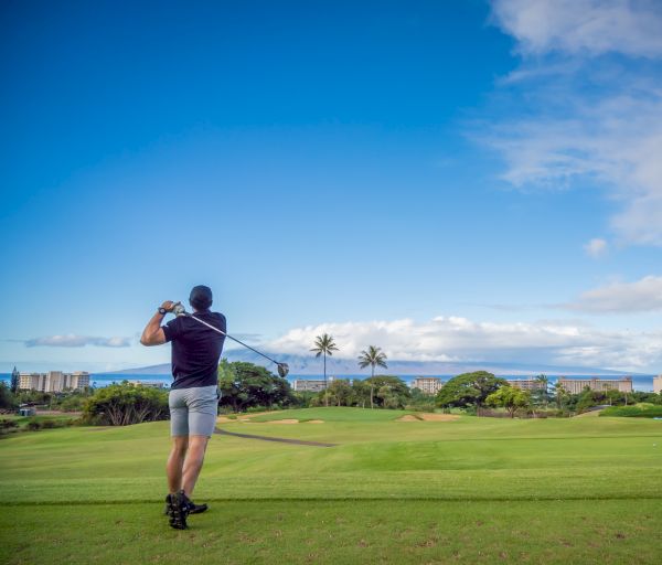 A man on a golf course swings a club, with a scenic background of greenery, palm trees, buildings, and a partly cloudy sky.