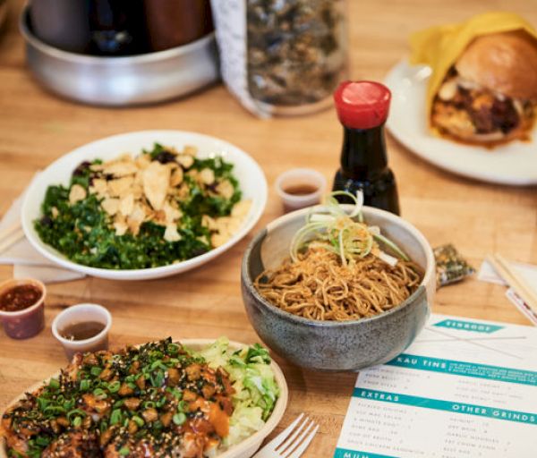 A table with various dishes including noodles, salad, a burger, a sauce bottle, and small dipping sauces, with a menu placed nearby.