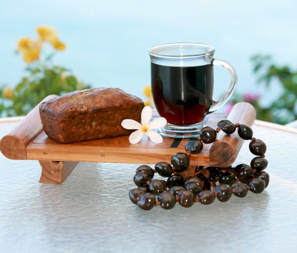 A wooden tray holds a loaf of bread, a glass mug of dark liquid, a flower, and a beaded necklace on a glass table with a scenic background.