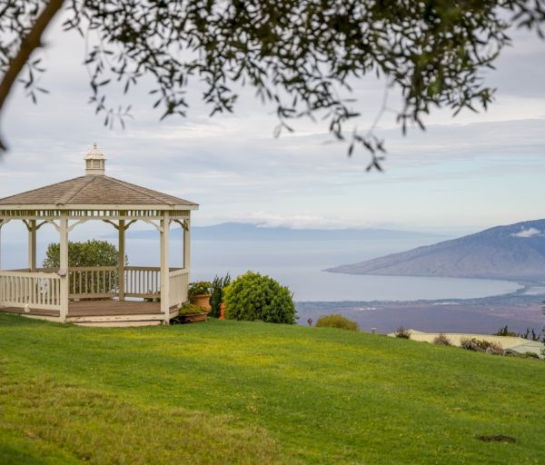 A white gazebo sits on a grassy hillside with a view of the ocean and distant hills under a cloudy sky.