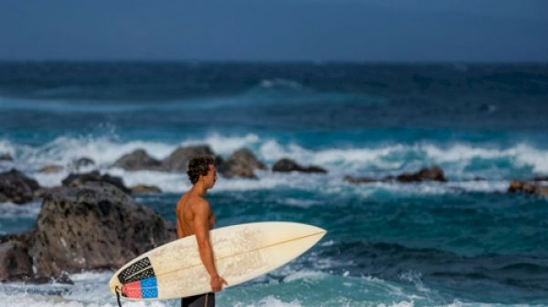 A person stands on the beach holding a surfboard, looking out at the ocean with rocks and waves in the background.