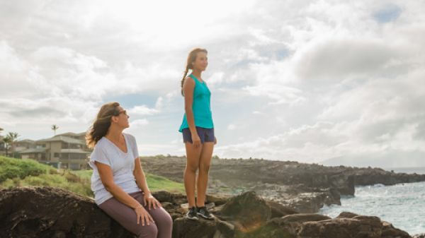 Two people, one sitting and one standing, are on rocky terrain near the ocean under a partly cloudy sky.