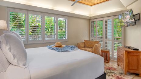 A cozy bedroom with a large bed, a hat and scarf on the bed, tropical-themed decor, wooden furniture, a TV, and windows with shutters.