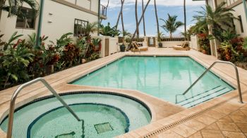 The image shows a tropical outdoor setting with a swimming pool and hot tub, surrounded by lush plants and palm trees, with buildings nearby.