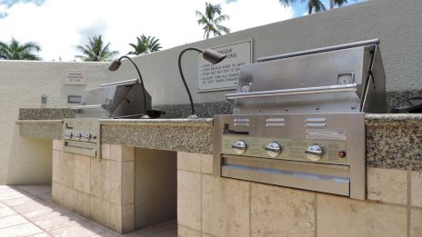 The image shows two stainless steel outdoor grills embedded in a stone countertop setup, with gooseneck reading lamps and a tropical background.