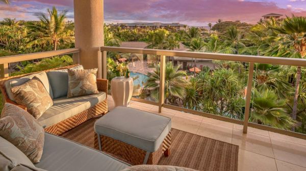 A balcony with comfortable seating overlooks a lush, green landscape and a swimming pool, with a vibrant sunset sky in the background.