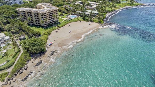 Aerial view of a beachfront with buildings, a sandy shore, and turquoise waters, surrounded by lush greenery and roads, ending at a rocky coastline.