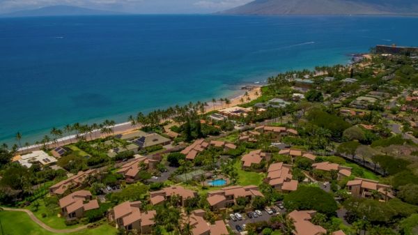 An aerial view of a coastal town with beachfront properties, palm trees, and the ocean leading to mountains in the background, on a sunny day.