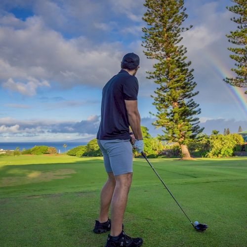 A person stands on a golf course, holding a club, looking at a rainbow in the distance with trees and a partly cloudy sky in the background.