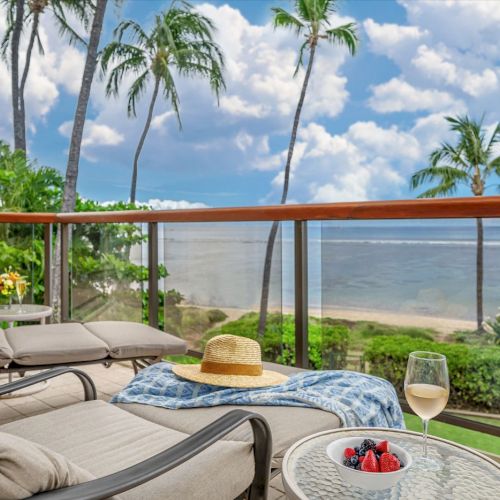 A relaxing patio with lounge chairs, a sun hat, fruit, and a drink on a table overlooks a tropical beach with palm trees and a bright sky.