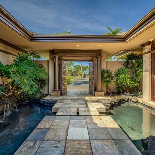 A beautiful entryway to a luxurious home, with a tiled path over water and lush greenery, leading to an open doorway and clear skies beyond.