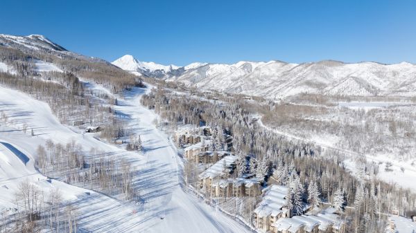 A snowy mountain landscape featuring ski slopes, a cluster of buildings, and trees, set against a backdrop of snow-covered peaks under a clear blue sky.