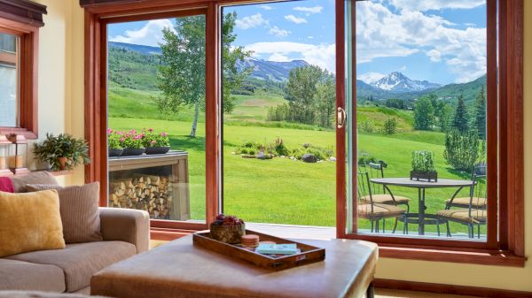 A cozy living room with large windows reveals a beautiful outdoor view of a green lawn, patio, trees, and distant mountains under a sunny sky.