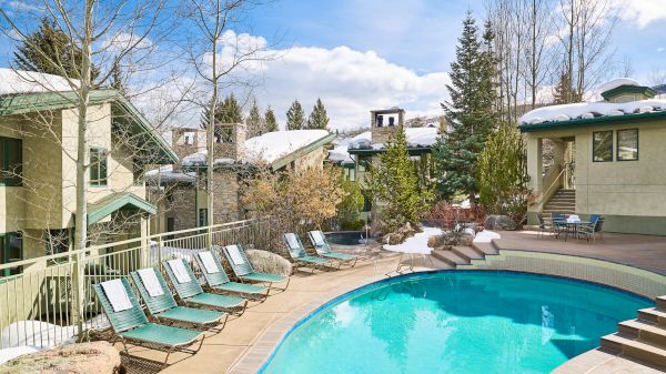 An outdoor pool area with several lounge chairs, surrounded by trees and buildings, under a clear blue sky with some clouds.