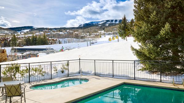 The image shows a pool and a hot tub on a patio overlooking a snow-covered landscape and ski slopes, with mountains in the background.