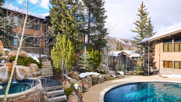 An outdoor area with a pool and hot tub surrounded by trees and buildings, with stairs leading down and light snow on the ground.