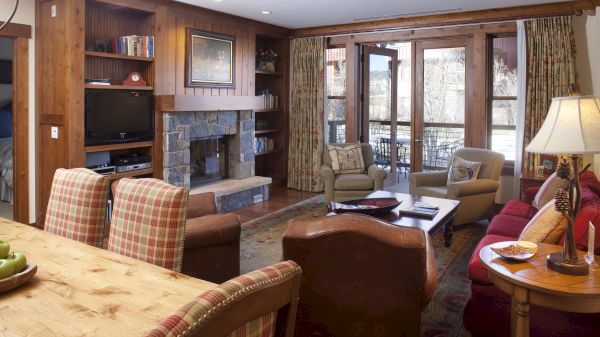 A cozy living room with a fireplace, TV, plaid chairs, and large windows leading to an outdoor patio area.