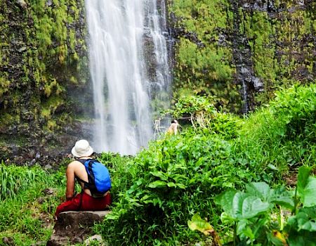 A person with a blue backpack and white hat sits on a rock, admiring a waterfall surrounded by lush green vegetation.