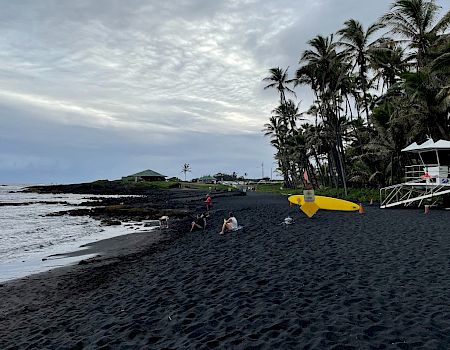 A beach with dark sand, a lifeguard tower, palm trees, and a few people near the water under a cloudy sky.