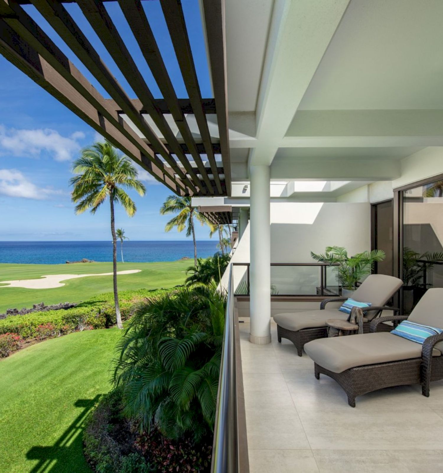 A patio with lounge chairs overlooks a golf course by the ocean, with palm trees and lush greenery in the background.