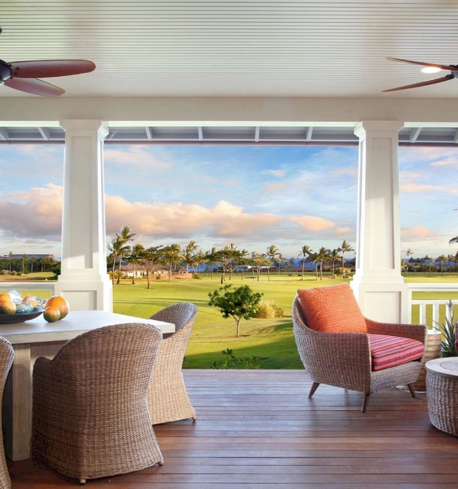 A cozy outdoor patio with wicker furniture, ceiling fans, and a view of a lush green lawn, palm trees, and a serene landscape.