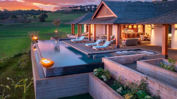 A luxurious modern villa with an infinity pool, lounge chairs, covered outdoor seating, lush green surroundings, and a beautiful sunset in the background.