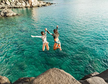 Two people are seen mid-air, jumping off rocks into clear turquoise water, amidst a rocky shoreline backdrop.