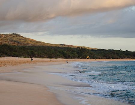 A scenic beach with calm waves, a few people relaxing on the sand, lush greenery, and hills under a partly cloudy sky.